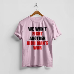 May include: A light pink t-shirt with the text "WE WON'T FIGHT ANOTHER RICH MAN'S WAR" in black and red lettering. The shirt is hanging on a wooden hanger against a white background.