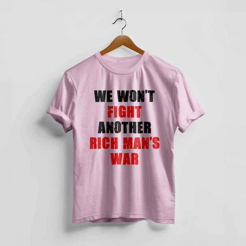 May include: A light pink t-shirt with the text "WE WON'T FIGHT ANOTHER RICH MAN'S WAR" in black and red lettering. The shirt is hanging on a wooden hanger against a white background.