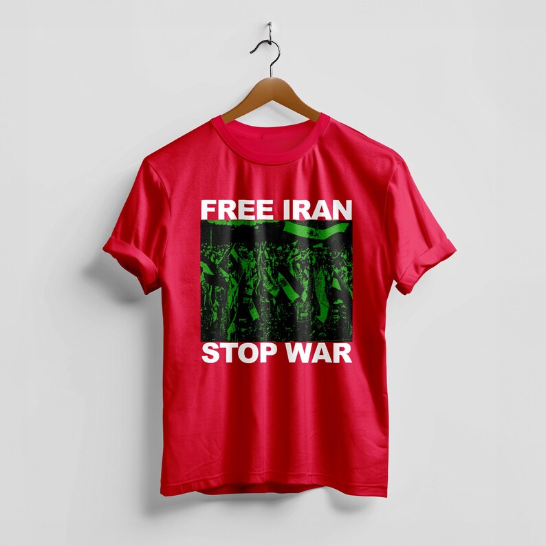 Op de afbeelding: Een rood t-shirt met de woorden "FREE IRAN" en "STOP WAR" in witte blokletters. Het shirt heeft een groen-zwarte afbeelding van een menigte met vlaggen. Het t-shirt hangt aan een houten hanger.