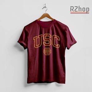 Op de afbeelding: Bordeauxrood t-shirt met de letters "USC" in goud, omlijnd in bordeaux. Daaronder een goud en bordeaux embleem. Het t-shirt hangt aan een houten hanger.