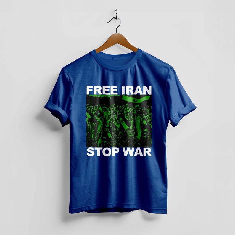 Op de afbeelding: Koningsblauw t-shirt met de witte tekst "FREE IRAN" en "STOP WAR". Het shirt heeft een groen-zwarte afbeelding van een menigte met vlaggen. Het t-shirt hangt aan een houten kleerhanger.