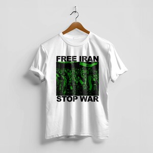 Op de afbeelding: Wit t-shirt met de zwarte tekst "FREE IRAN STOP WAR". Het shirt heeft een groen-zwarte afbeelding van een menigte met vlaggen. Het t-shirt hangt aan een houten hanger.