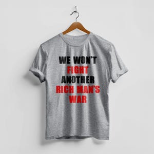 May include: A heather gray t-shirt with the text "WE WON'T FIGHT ANOTHER RICH MAN'S WAR" in black and red block letters. The shirt is hanging on a wooden hanger against a white background.