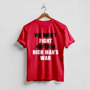 May include: A red t-shirt with the text "WE WON'T FIGHT ANOTHER RICH MAN'S WAR" in a mix of black and white fonts. The shirt is hanging on a wooden hanger against a white background.