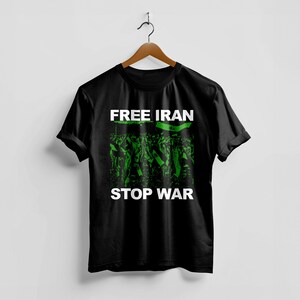 Op de afbeelding: Zwart T-shirt met de woorden "FREE IRAN STOP WAR" in witte blokletters. Het shirt heeft een groen-zwarte afbeelding van een menigte die vlaggen vasthoudt. Het shirt hangt aan een houten hanger.