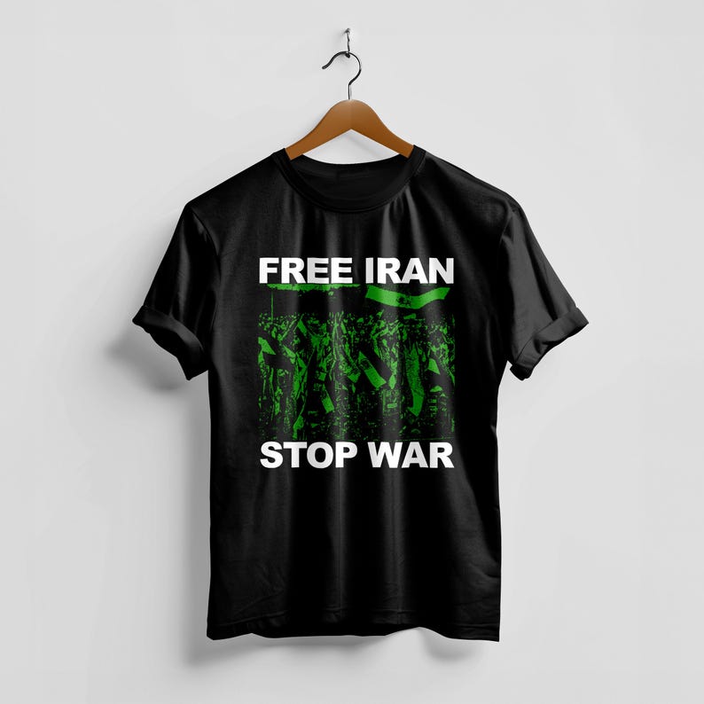 Op de afbeelding: Zwart T-shirt met de woorden "FREE IRAN STOP WAR" in witte blokletters. Het shirt heeft een groen-zwarte afbeelding van een menigte die vlaggen vasthoudt. Het shirt hangt aan een houten hanger.