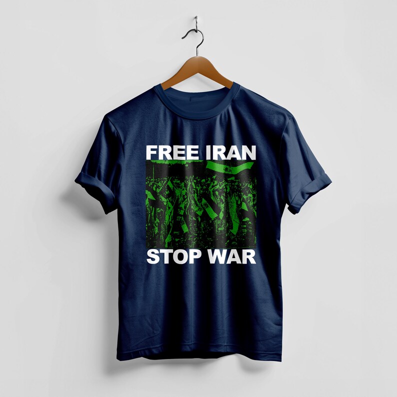 Op de afbeelding: Marineblauw t-shirt met de witte tekst "FREE IRAN STOP WAR". De afbeelding in het midden toont een groen-zwarte afbeelding van een menigte die vlaggen vasthoudt. Het t-shirt hangt aan een houten hanger.