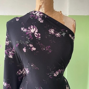May include: A length of black fabric draped over a dress form, featuring a repeating floral pattern in shades of purple, white, and muted green. The material has a smooth, slightly reflective surface and is arranged in an asymmetrical style.