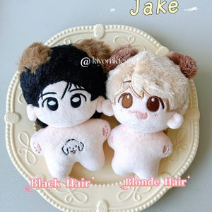 May include: Two plush dolls with cartoon faces and fluffy hair, one with black hair and the other with blonde hair, are displayed on a decorative plate. The dolls have pink bodies and embroidered facial features. Text on the plate reads "Black Hair" and "Blonde Hair".