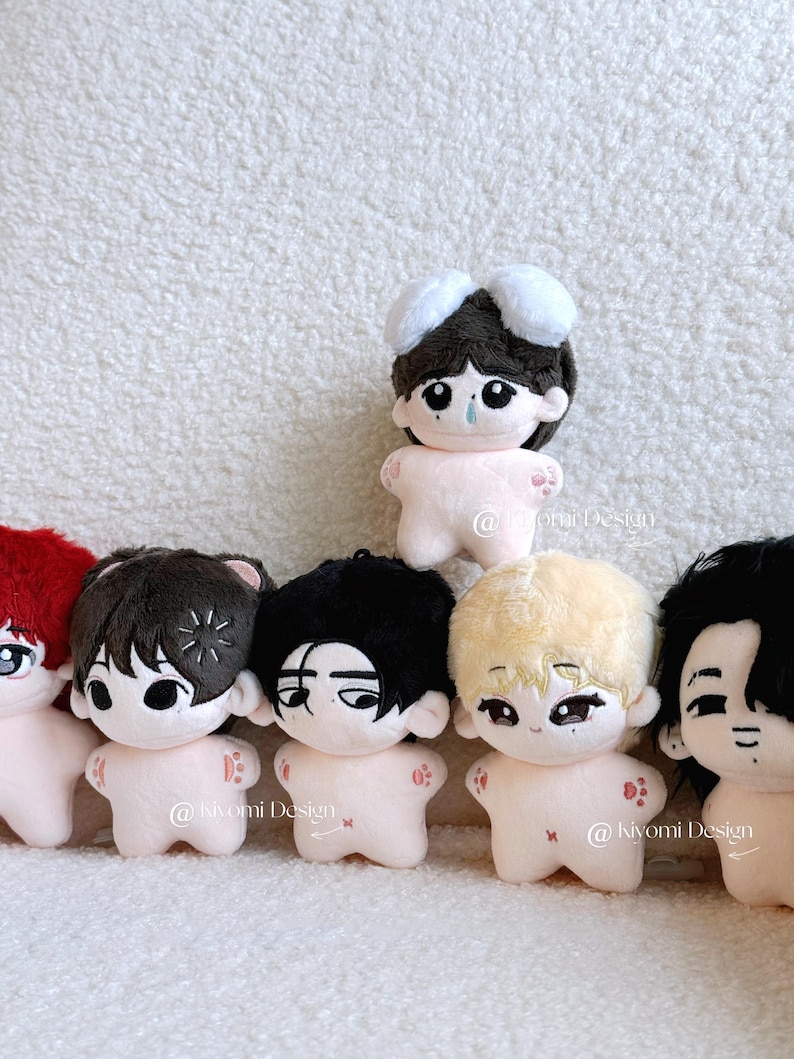 May include: A collection of small plush dolls with various hairstyles and facial expressions. The dolls have light pink bodies and detailed embroidered faces. The dolls have different hair colors, including black, brown, and blonde.