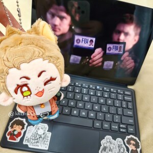May include: A plush doll with a cartoon character design, featuring a light brown fur-like texture and a smiling face. The doll is positioned in front of a laptop with stickers and a screen displaying a scene with the text "FBI". The doll is attached to a silver chain.