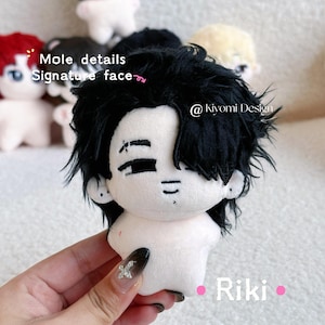 May include: A plush doll with black hair and a signature face, held in a hand. The doll has a white body and is surrounded by other similar dolls. The text "Mole details Signature face" is visible.