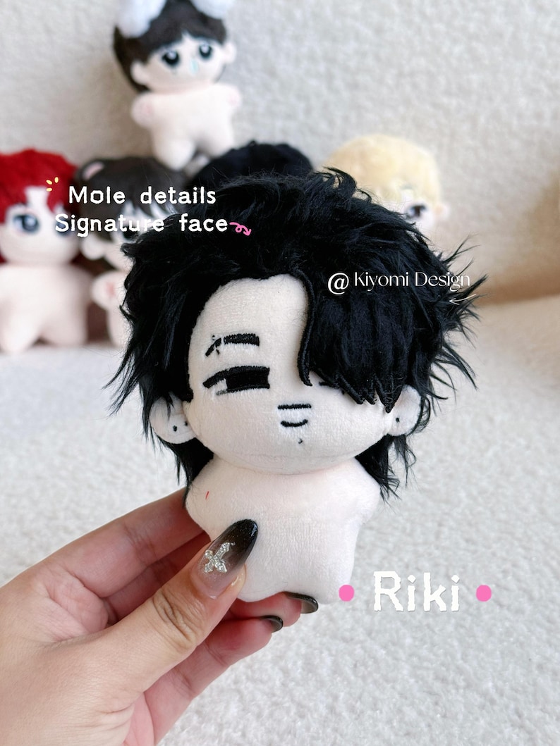 May include: A plush doll with black hair and a signature face, held in a hand. The doll has a white body and is surrounded by other similar dolls. The text "Mole details Signature face" is visible.