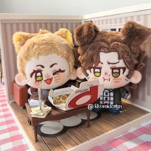 May include: Two plush dolls with anime-style faces, one with light brown hair and the other with dark brown hair, are seated in a miniature room. A small table holds a pizza box and a pizza. The dolls are in a miniature room setting.