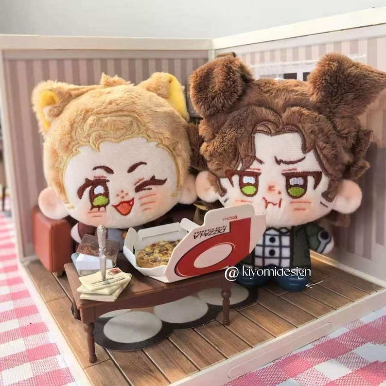 May include: Two plush dolls with anime-style faces, one with light brown hair and the other with dark brown hair, are seated in a miniature room. A small table holds a pizza box and a pizza. The dolls are in a miniature room setting.