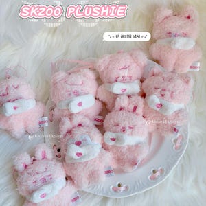 May include: Pink plushie toys with white accents and the text "SKZOO PLUSHIE". The toys are shaped like bears and bunnies, with closed eyes and small tags. The toys are on a white plate.