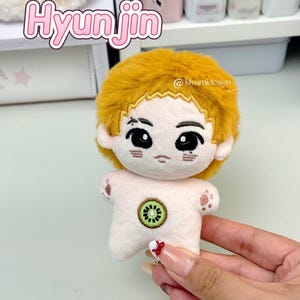 May include: A plush doll with a cartoon-like face and yellow hair. The doll has a kiwi fruit design on its torso and small paw prints on its arms. The name "Hyunjin" is written in pink bubble letters above the doll.