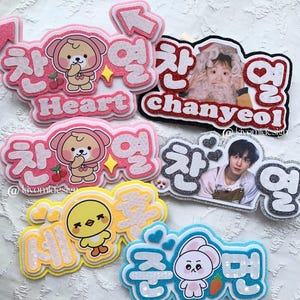 Peut inclure: Plusieurs badges nominatifs scintillants avec des personnages de dessins animés et du texte coréen. Les motifs incluent un ours, un poussin et un lapin, avec le nom "Chanyeol" et le mot "Heart" en anglais. Les badges sont dans des tons de rose, jaune et bleu.