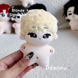 May include: A plush doll with blonde hair and a signature smile. The doll has large eyes and small red paw prints on its arms. The text "Blonde hair Signature smile" is visible in the image. The doll is held by a person.