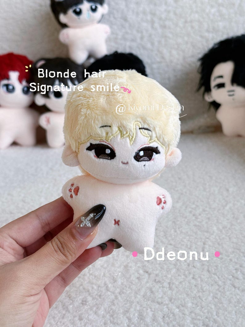 May include: A plush doll with blonde hair and a signature smile. The doll has large eyes and small red paw prints on its arms. The text "Blonde hair Signature smile" is visible in the image. The doll is held by a person.