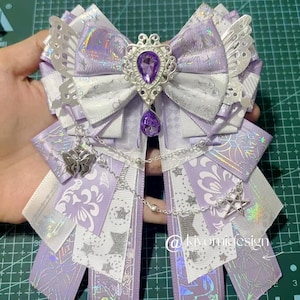 May include: A decorative bow in lavender and white, with detailed ribbon work and embellishments. The bow features a large central jewel and dangling charms. The design includes star and floral patterns, with the text "@kivomidesign".