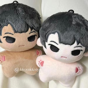 May include: Two plush dolls with gray hair and stern facial expressions. One doll is tan, the other is light pink. Both have small red paw prints on their feet. The dolls have a black loop at the top for hanging.