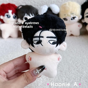 May include: A plush doll with black hair, signature eyebrows, and mole details. The doll has a light skin tone and is holding a hand. The text "Hoonie A" and "@kiyomi Design" are visible.