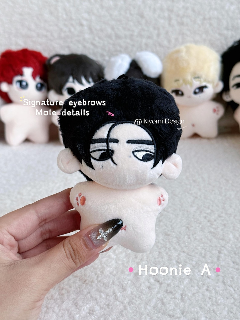 May include: A plush doll with black hair, signature eyebrows, and mole details. The doll has a light skin tone and is holding a hand. The text "Hoonie A" and "@kiyomi Design" are visible.