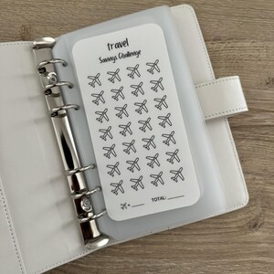 May include: White travel savings challenge tracker with a grid of 40 airplane icons. The tracker has a space to write the total amount saved.