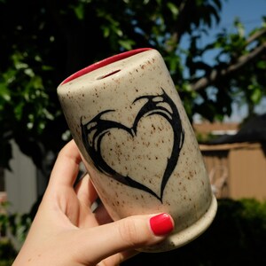 May include: A speckled ceramic mug with a black heart design. The mug has a red interior and is held against a backdrop of green foliage and a blue sky. The heart design is stylized and abstract.
