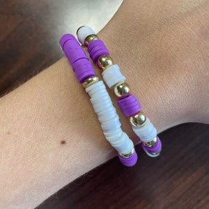 May include: Two bracelets with white and purple beads and gold accents. The bracelets are stacked on a person's wrist.