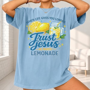 May include: A light blue t-shirt featuring a graphic print of a yellow lemon, a lemon slice, and a glass jar of lemonade. The text reads: WHEN LIFE GIVES YOU LEMONS TRUST JESUS HE MAKES LEMONADE. The design includes small yellow star accents.