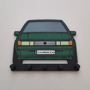 Puede incluir: Un llavero Volkswagen Scirocco verde con una base negra y cinco ganchos. El coche está representado en un estilo de dibujos animados con una matrícula que dice "Scirocco".