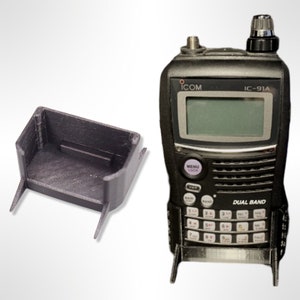 以下が含まれることがあります： 黒色のICOM IC-91Aデュアルバンド無線機と、それに合わせた黒色の3Dプリントスタンド。無線機には画面、ボタン、アンテナがあります。スタンドは無線機を直立させるように設計されています。無線機の高さは約15cmです。