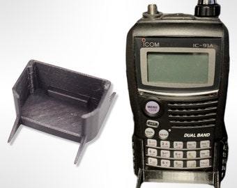 Pode incluir: Um rádio dual band ICOM IC-91A preto com um suporte preto impresso em 3D a condizer. O rádio tem um ecrã, botões e uma antena. O suporte foi concebido para manter o rádio na posição vertical. O rádio tem aproximadamente 15 cm de altura.
