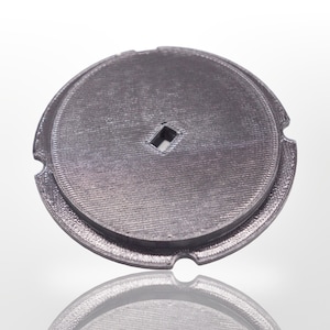 May include: A circular, metallic gray object with a textured surface and a square hole in the center. The object has a raised outer rim and two curved cutouts. The object is likely a component or part of a larger mechanism.