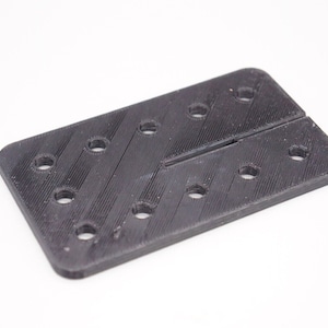 May include: A black plastic rectangular plate with multiple holes and a thin slot in the center.