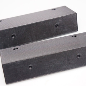 May include: Two black rectangular blocks with rounded edges and several drilled holes. The blocks are made of a textured material and are positioned on a white surface.
