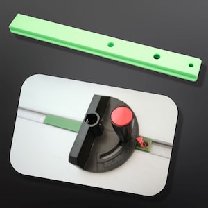 May include: A green plastic guide rail with three holes and a black and red circular tool with a knob and a scale. The tool is attached to a white surface.