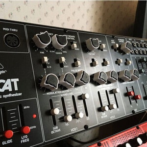Könnte beinhalten: Ein schwarzer Behringer CAT elektronischer Musiksynthesizer mit Knöpfen und Schiebereglern. Der Synthesizer hat ein weißes Katzenlogo und den Text "CAT" in weißen Buchstaben.