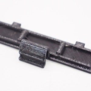 May include: A black 3D-printed object, possibly a component or part, with a rectangular base and a cylindrical piece attached. The object has a textured surface and is set against a white background.