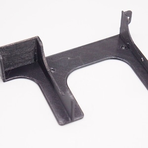 May include: A black plastic 3D printed bracket with two open slots. The bracket has a curved shape and is designed to hold something in place.