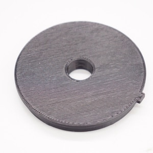 Puede incluir: Un disco de filtro de plástico negro con un agujero central. El disco tiene una superficie texturizada y una forma ligeramente irregular.