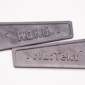 Peut inclure: Deux plaques de nom en plastique gris avec des coins arrondis. La plaque supérieure porte le mot "KORG" en relief. La plaque inférieure porte le texte "Nu: Tek" en relief.