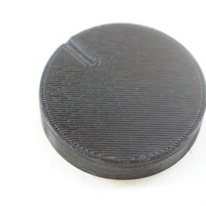 Può includere: Un oggetto rotondo nero stampato in 3D con una superficie testurizzata e una piccola scanalatura sul lato.