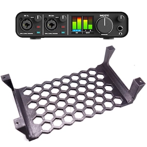 May include: Black metal rack with a honeycomb design, designed to hold a small audio interface. The interface is black with a green and red display and the text "MOTU" on the front.