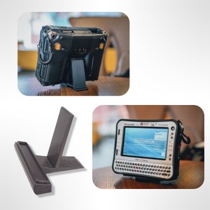 May include: A black and grey ruggedised tablet computer with a stand. The tablet has a screen that says "TOUGHBOOK" and is on a wooden table. The stand is made of black plastic and has a textured surface.