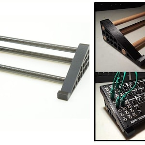 Może przedstawiać: Czarny stojak 3D z trzema prętami z włókna węglowego do syntezatora. Stojak jest pochylony, aby podnieść syntezator. Stojak jest pokazany z czarnym syntezatorem z napisem "Moog" na nim.