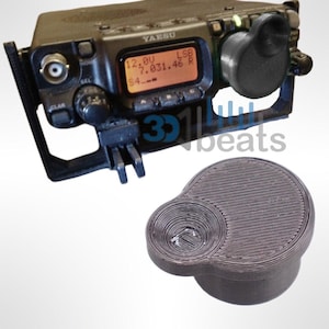May include: A black and gray 3D printed microphone for a radio. The microphone has a round, flat base and a smaller, round, raised section in the center. The microphone is designed to fit on a radio.