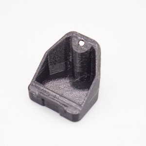 May include: A black, 3D-printed, angled holder with a rectangular base and a small hole at the top. The holder has a textured surface and is designed to hold a small object. The item is made of plastic.